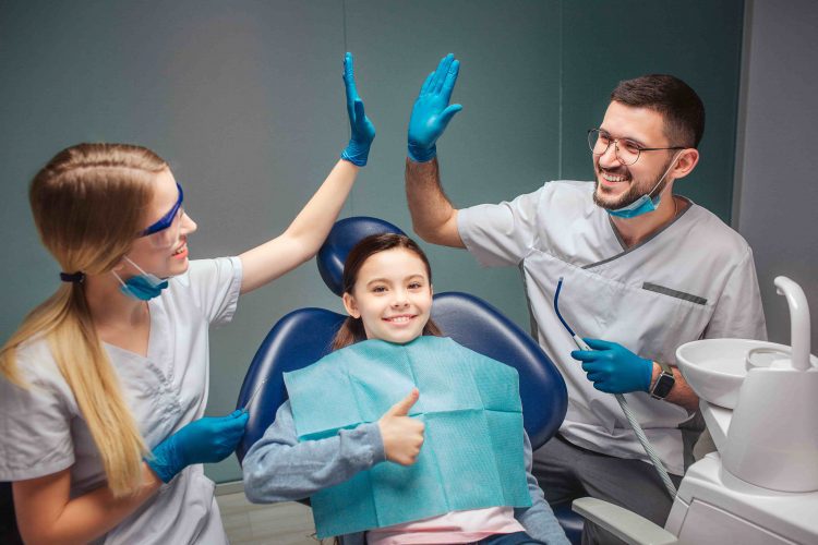 Dentistas sorrindo e se cumprimentando enquanto menina com mordida cruzada sorri na cadeira