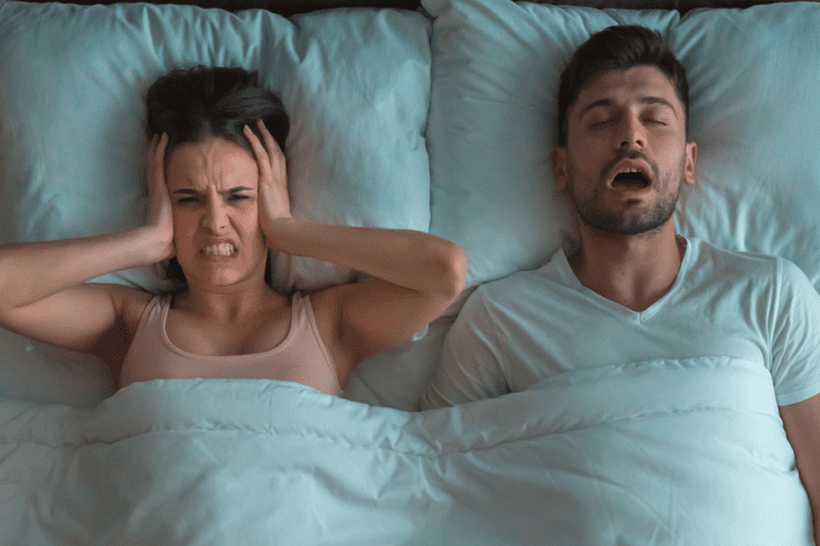 Casal na cama: homem roncando e mulher incomodada, destacando os impactos e o que é apneia do sono no dia a dia.