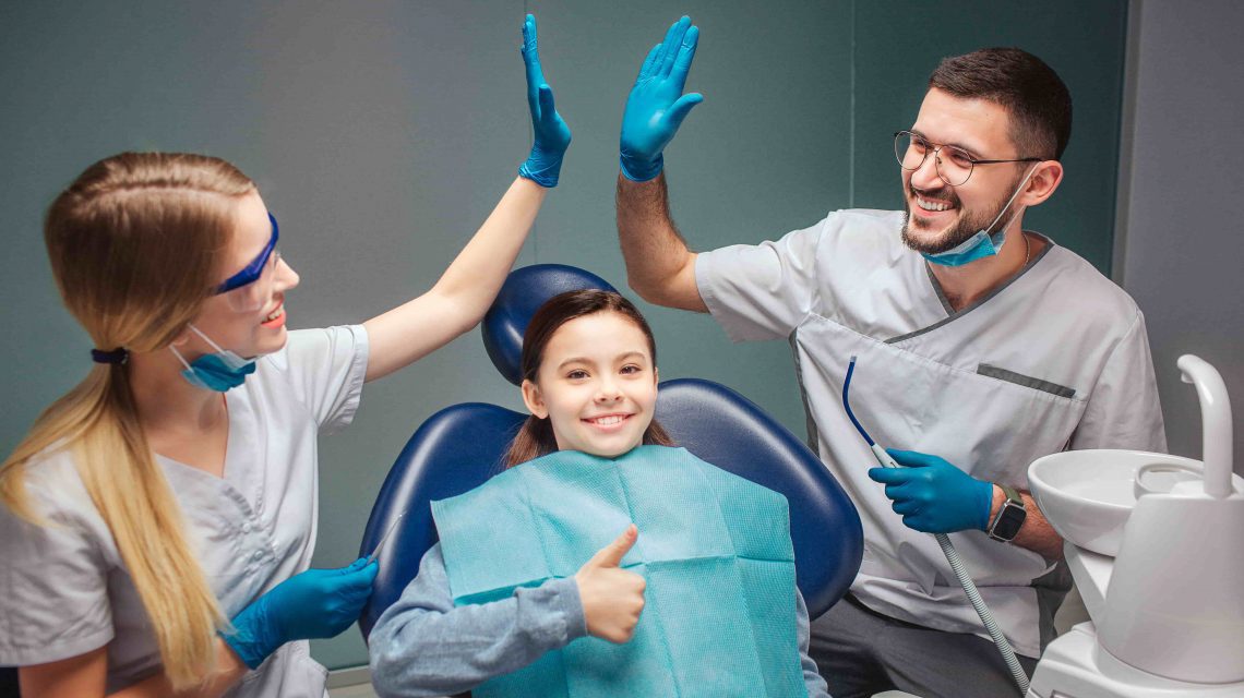 Dentistas sorrindo e se cumprimentando enquanto menina com mordida cruzada sorri na cadeira