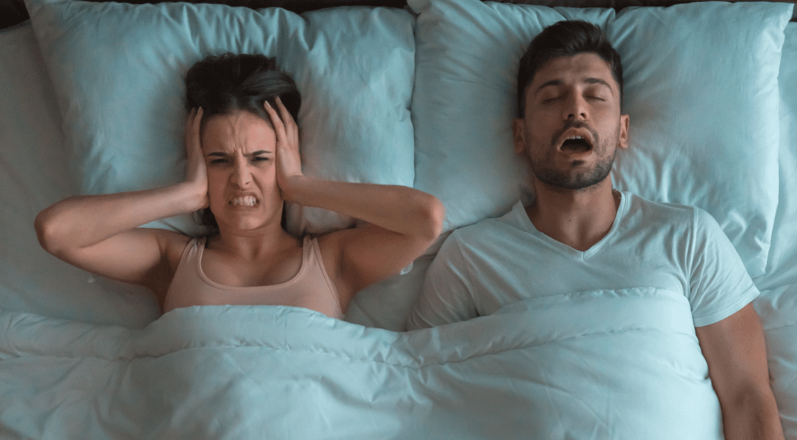 Casal na cama: homem roncando e mulher incomodada, destacando os impactos e o que é apneia do sono no dia a dia.