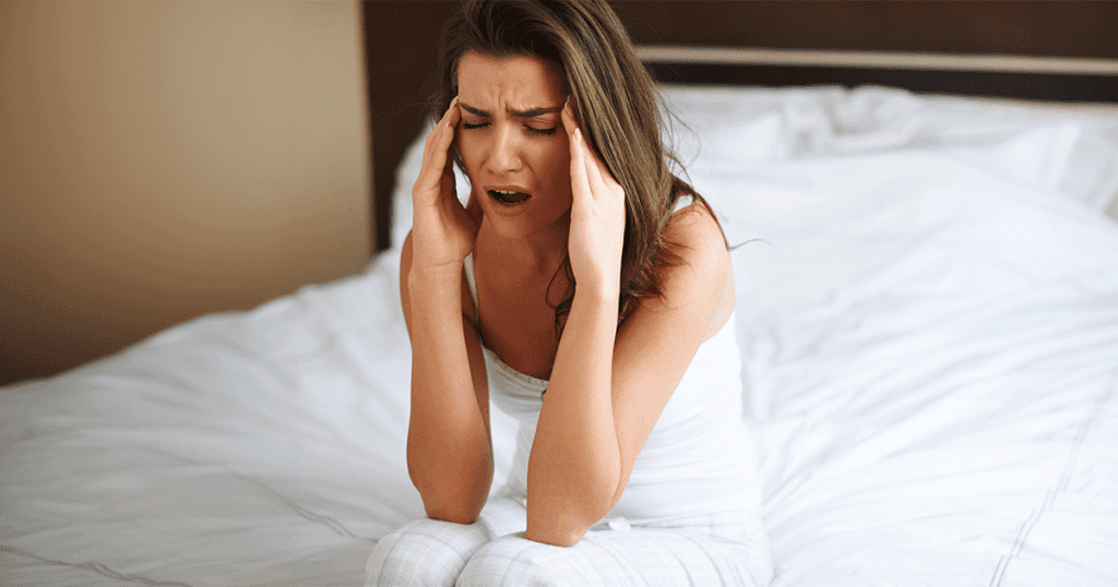 Mulher sentada na cama com expressão de cansaço e dor de cabeça, ilustrando sintomas de o que é apneia do sono e suas consequências.